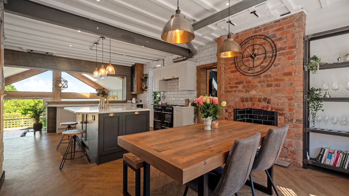 An open kitchen and dining area showcases rustic charm with exposed brick walls and wood beams. A large wooden dining table is surrounded by gray upholstered chairs, while modern pendant lights hang above. A kitchen island, equipped with bar seating, complements the space.