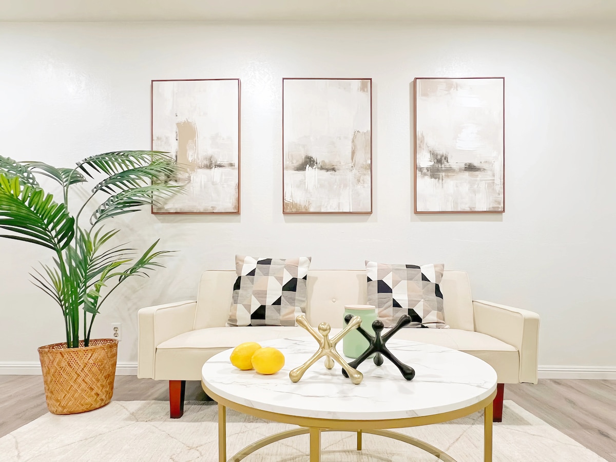 A modern living area showcases a light-colored sofa adorned with patterned cushions. A round coffee table displays decorative items, including fruit and unique sculptures. Three framed artworks hang on the wall, while a potted plant adds a touch of greenery to the space.
