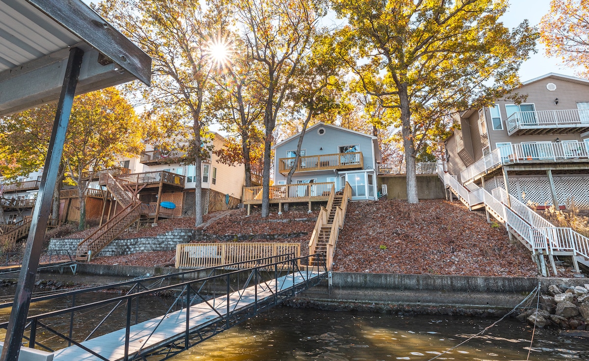 A newly remodeled home is shown from the water, surrounded by trees with autumn leaves. A private dock extends towards the house, which features multiple levels and outdoor stairs leading to a spacious deck. Sunlight filters through the trees, creating a serene ambiance.