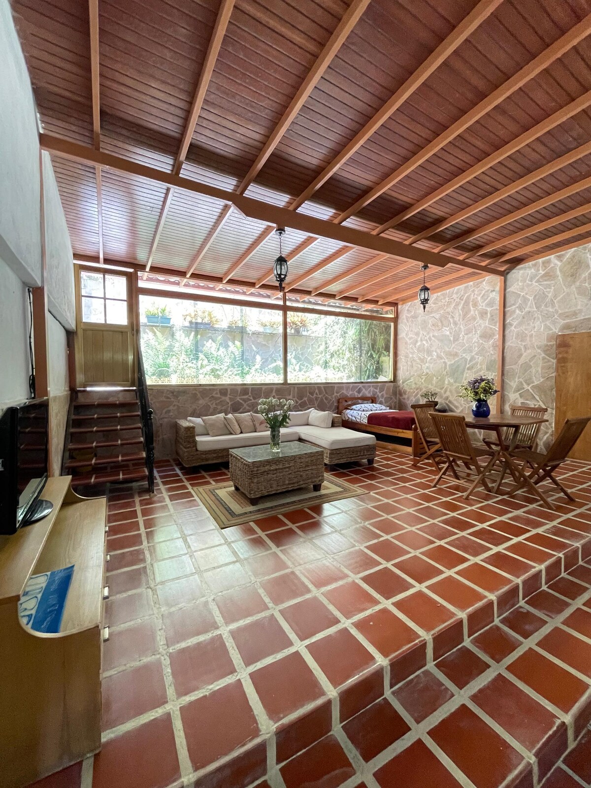 A cozy living area features a combination of natural stone walls and a decorative tiled floor. Comfortable seating arrangements include a spacious sofa and a coffee table with a floral centerpiece. A dining table is set near a large window, allowing plenty of natural light.