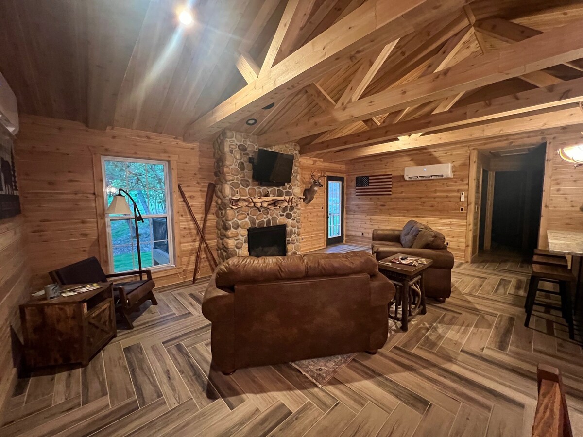 An inviting living area features a stone fireplace as the focal point, with a large flat-screen television mounted above. Comfortable seating includes a brown leather sofa and armchair, complemented by wooden side tables. Natural light enters through large windows, enhancing the wooden interior design.