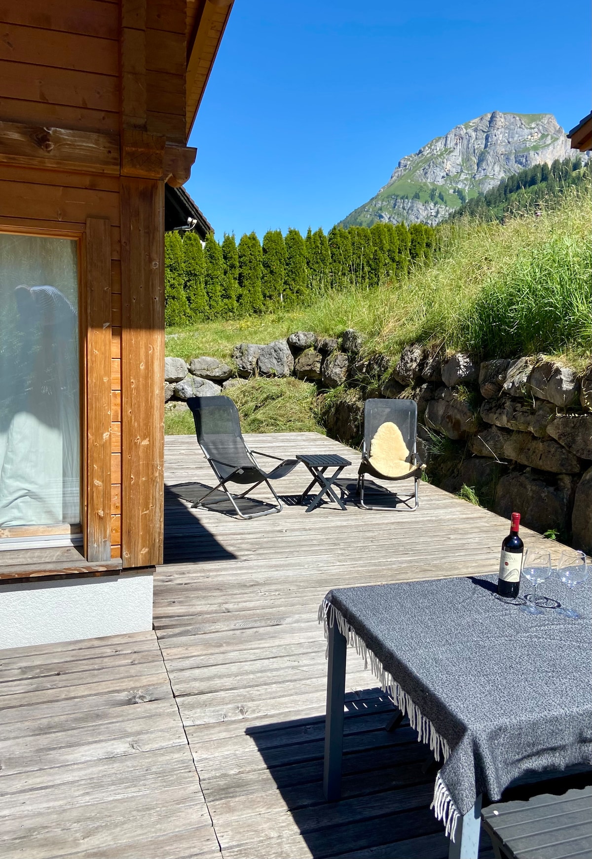A wooden terrace is displayed with a table covered by a dark tablecloth and wine glasses set for enjoyment. Two lounge chairs are positioned nearby, allowing for relaxation in the sun. Lush greenery and a mountain backdrop create a serene environment.