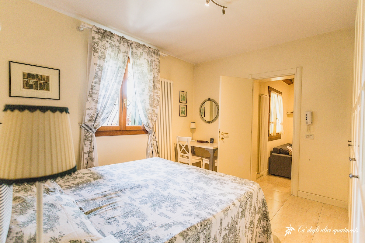 A double bedroom features light-colored walls adorned with framed artwork. A spacious bed is covered with a patterned bedspread, and a large window with floral curtains allows natural light. A small desk and chair are positioned in the corner, enhancing the room's functionality.
