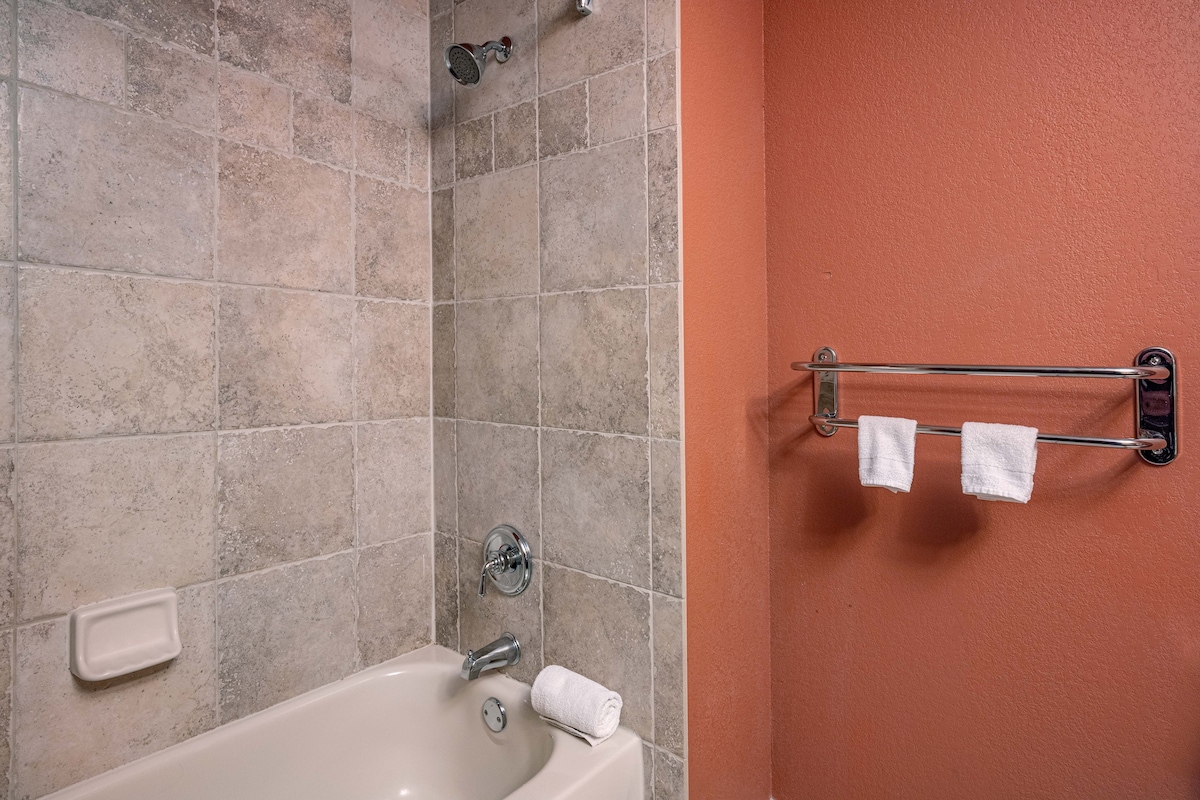 A full bathroom is shown, featuring a bathtub with a handheld shower. The walls are adorned with neutral tile and an accent painted in warm tones. A towel rack with two white towels is mounted nearby, adding functionality to the space.
