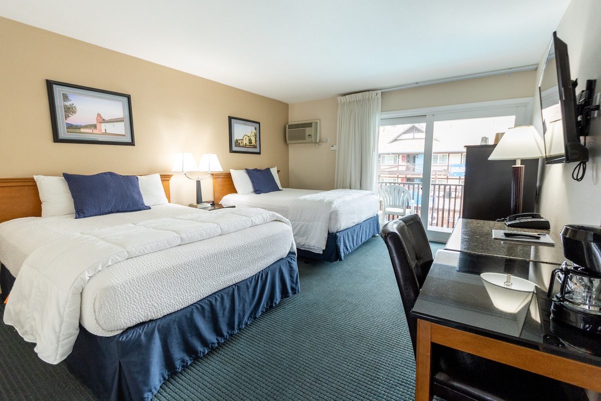 A spacious room features two beds with white linens and decorative dark blue accents. A desk and a coffee maker are positioned near a large window that offers natural light. Art pieces adorn the walls, complementing the room's warm color palette.