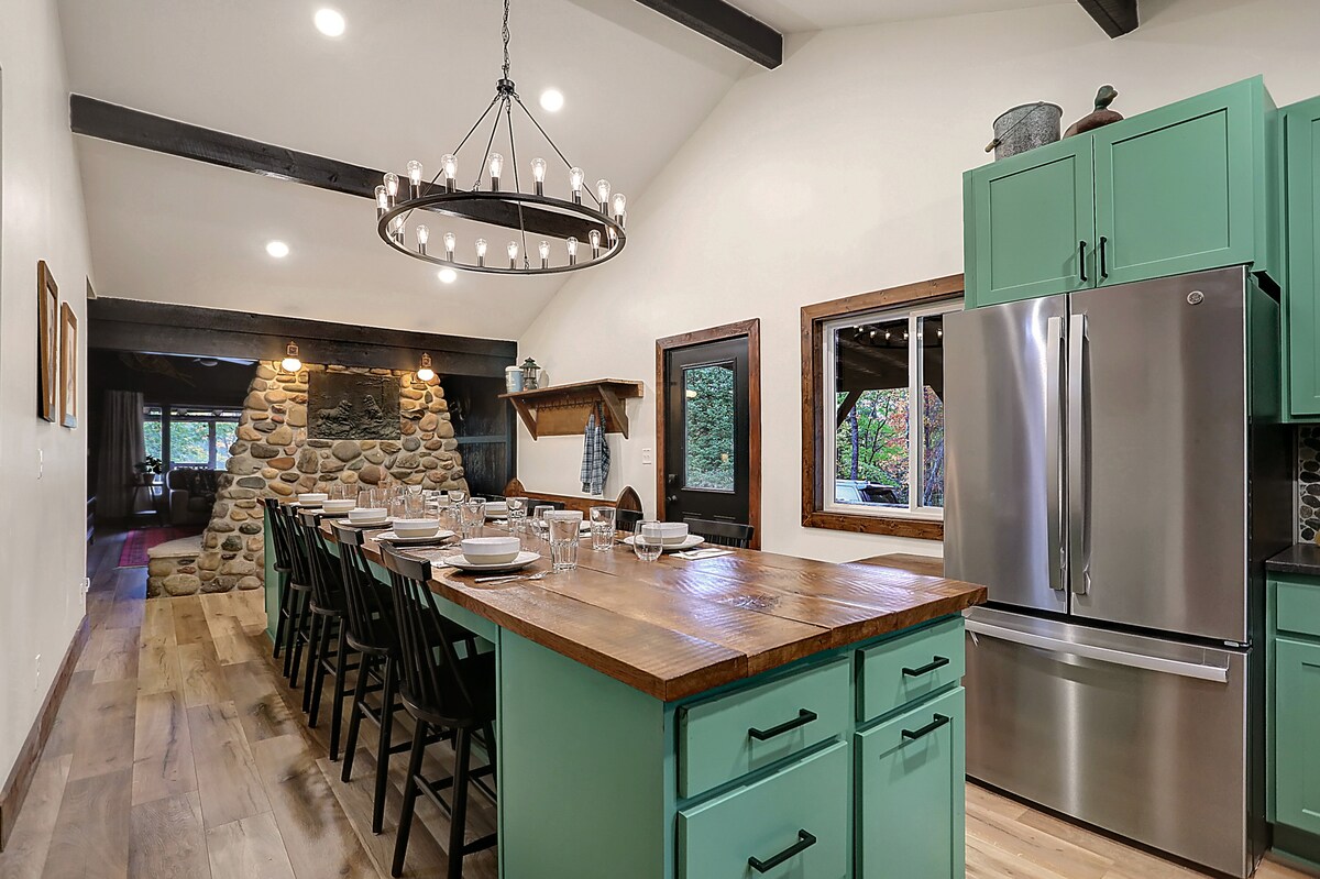 The spacious kitchen features a custom dining table island with ample seating. A large stone fireplace is positioned in the background, creating a lodge-like ambiance. Modern appliances, including a stainless steel refrigerator, are present, with windows allowing natural light to brighten the space.