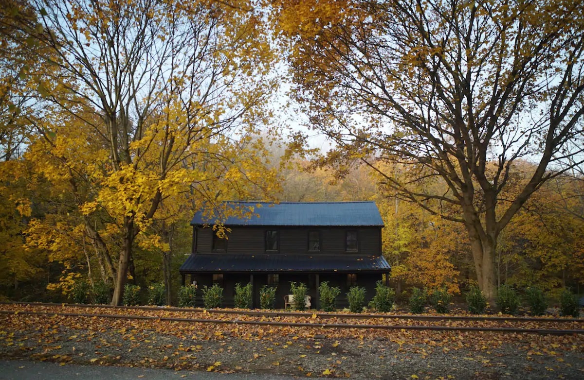 The exterior of a two-story cabin is framed by vibrant autumn foliage. A dark wooden facade contrasts with the bright yellow and orange leaves, while the structure is set back from the Northern Central Railway Trail. The surrounding landscape features a mix of trees and shrubs.