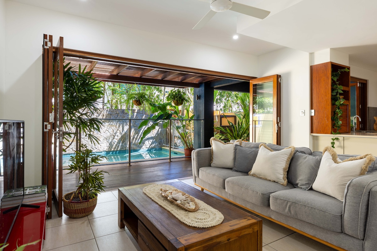 An open living space features a gray sofa arranged around a wooden coffee table. Large sliding doors provide a view of a private pool surrounded by greenery. Potted plants contribute to the tropical atmosphere, while natural light fills the area.