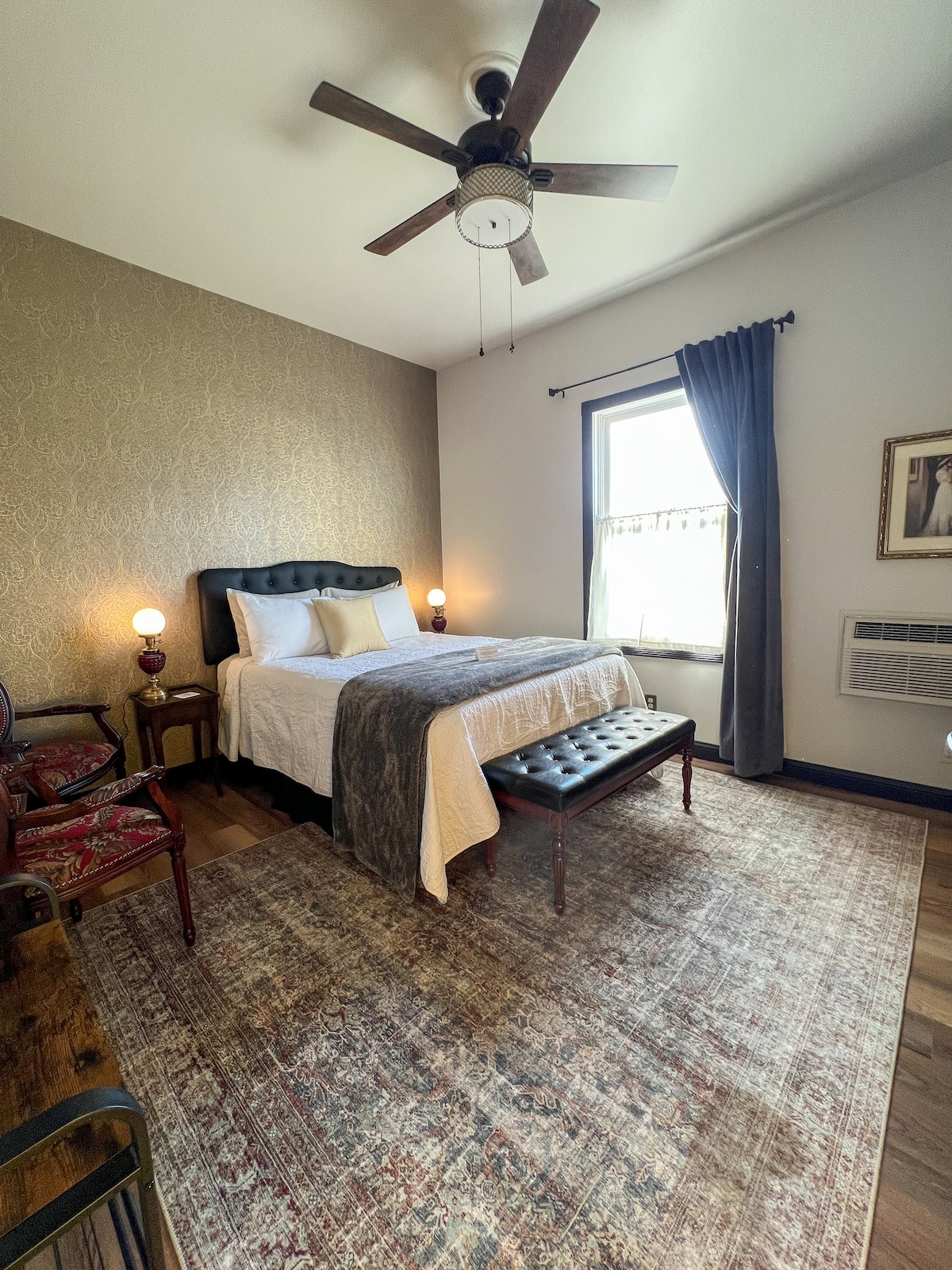 A serene room is presented with a comfortable queen-size bed, accompanied by a decorative bench at its foot. Warm light is provided by bedside lamps, while a large window is dressed with dark curtains. A partial view of the ornate wallpaper and wooden flooring is visible.