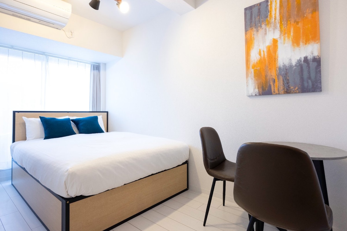 A spacious sleeping area features a large bed with white linens and accent pillows in shades of blue. A circular table and two chairs provide a cozy dining space. A colorful abstract painting adorns the wall, and natural light enters through a large window.