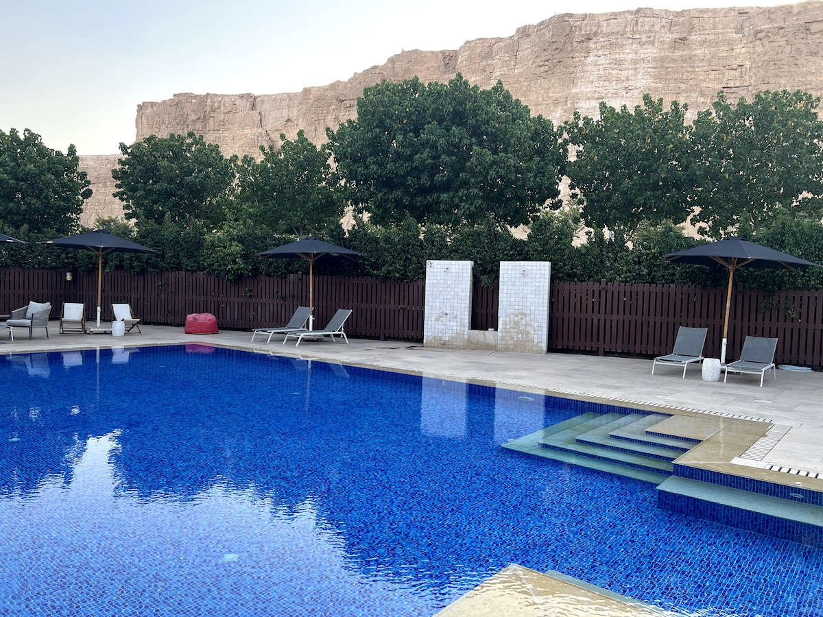 An outdoor pool area is highlighted by a sparkling blue surface and surrounded by comfortable lounge chairs. Umbrellas provide shaded spots, while lush greenery and a rustic wooden fence create a serene backdrop against the rugged rock formations in the distance.