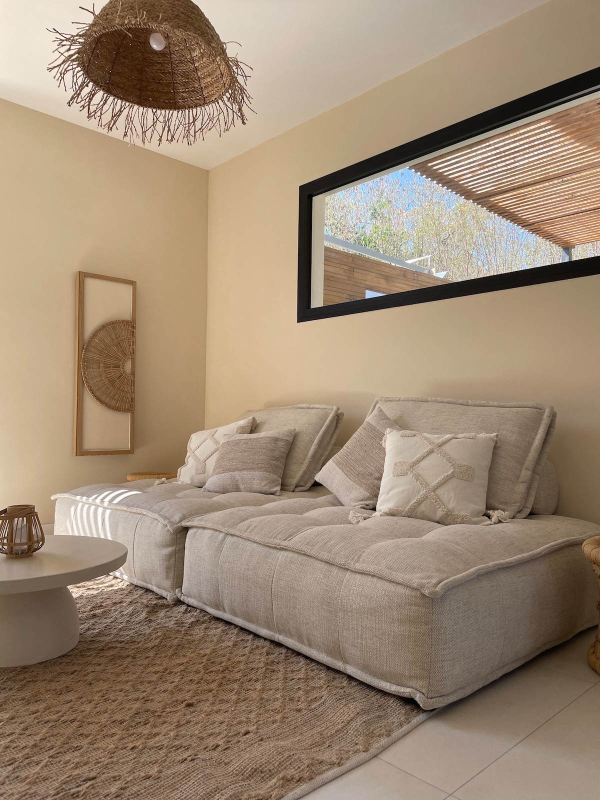 A cozy seating area is present, featuring a light-colored, plush couch with decorative cushions. A round coffee table rests on a textured area rug, while natural light streams in through a large window, highlighting the warm tones of the walls.