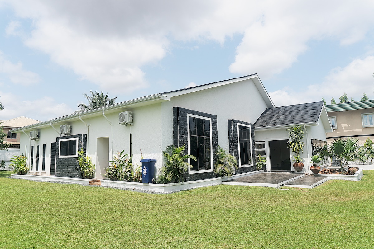 A modern villa is showcased, featuring a white exterior with black stone accents. Large windows enhance natural light, while a well-kept lawn surrounds the building. Potted plants add greenery, contributing to the inviting outdoor space. The driveway is visible, leading to an entrance with overhang.