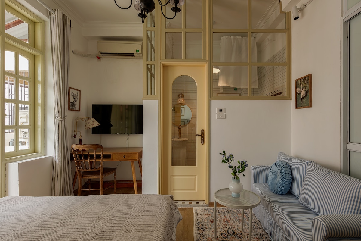 A compact living space is defined by natural light from large windows and a neutral color palette. A comfortable bed is paired with a cozy sofa, while a wooden desk is positioned near a TV. A glass door leads to a private bathroom, enhancing the layout.