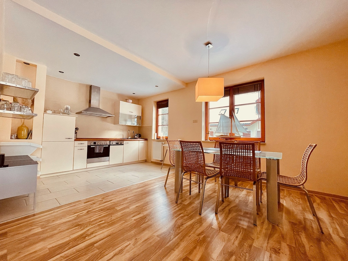 A modern kitchen and dining area features sleek cabinetry and stainless steel appliances. A glass dining table is surrounded by four woven chairs. Natural light fills the space through large windows, highlighting the wooden flooring and neutral wall tones.