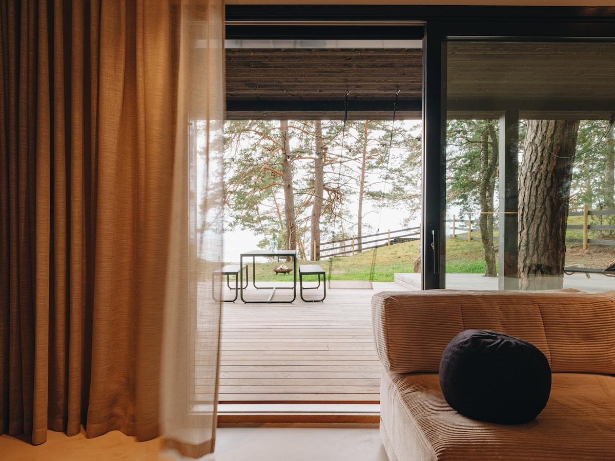 A spacious living area is visible through large glass doors, leading to a wooden deck furnished with a table and chairs. Soft curtains frame the view, while natural light floods the interior, enhancing the connection to the surrounding landscape and trees.