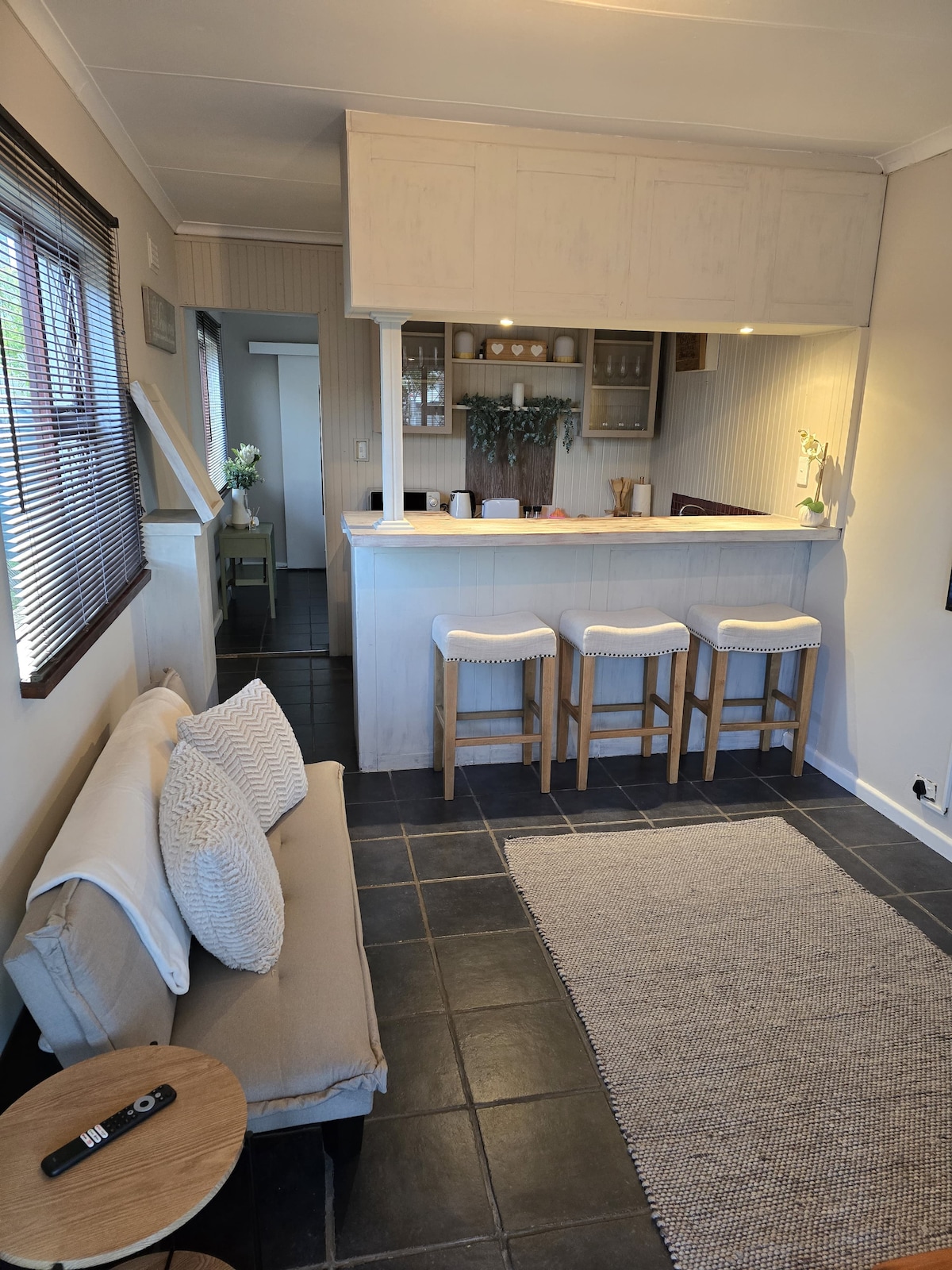The lounge area features a light-colored couch and a small wooden side table, complemented by a textured rug on the tiled floor. A compact kitchen counter showcases bar stools, with shelves above displaying kitchen essentials, creating an open and inviting atmosphere.