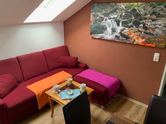 A cozy seating area is composed of a red couch with colorful throws in orange and purple. A wooden coffee table is placed in front, accompanied by two cups and a small plate. A large nature-themed artwork adorns the wall, adding visual interest.