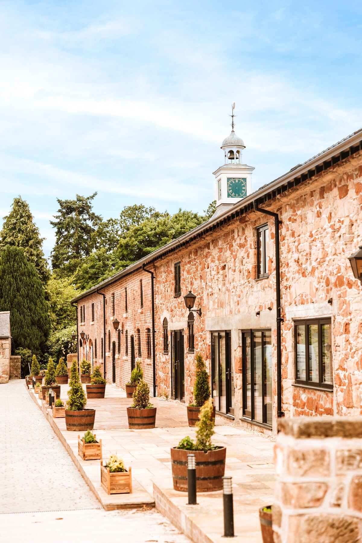 The exterior features a rustic stone building with large glass doors and windows reflecting the landscaped surroundings. Decorative planters with small trees line the pathway, adding greenery. A clock tower is visible in the background, contributing to the charming countryside setting.