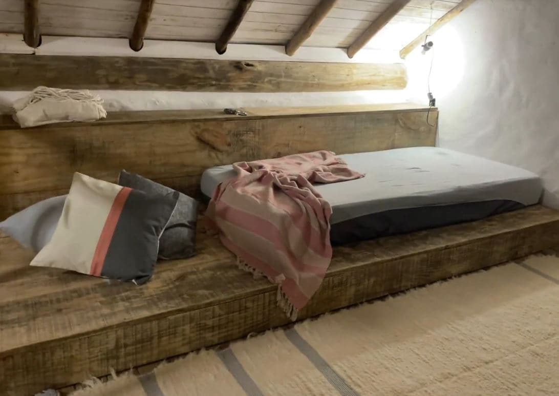 A cozy sleeping area features a raised wooden platform with a single mattress and soft blankets. Decorative pillows are arranged nearby. Natural wood beams are visible above, while a soft rug lies on the floor, contributing to a warm and rustic feel.