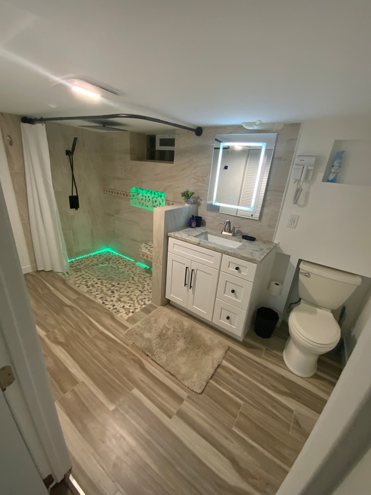 The bathroom features a modern design with warm-toned tiles and a spacious shower area. LED lighting adds a soft glow to the space. A white vanity with storage is illuminated by a stylish mirror. A toilet is positioned to one side, creating a functional layout.