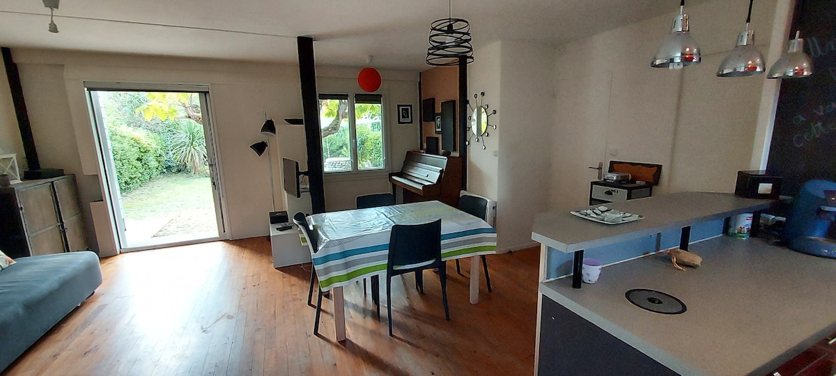 An open living space features a dining table with a striped tablecloth and black chairs. A well-equipped kitchen and a piano are included. Large windows allow natural light to illuminate the area, and a door leads to a garden visible outside.