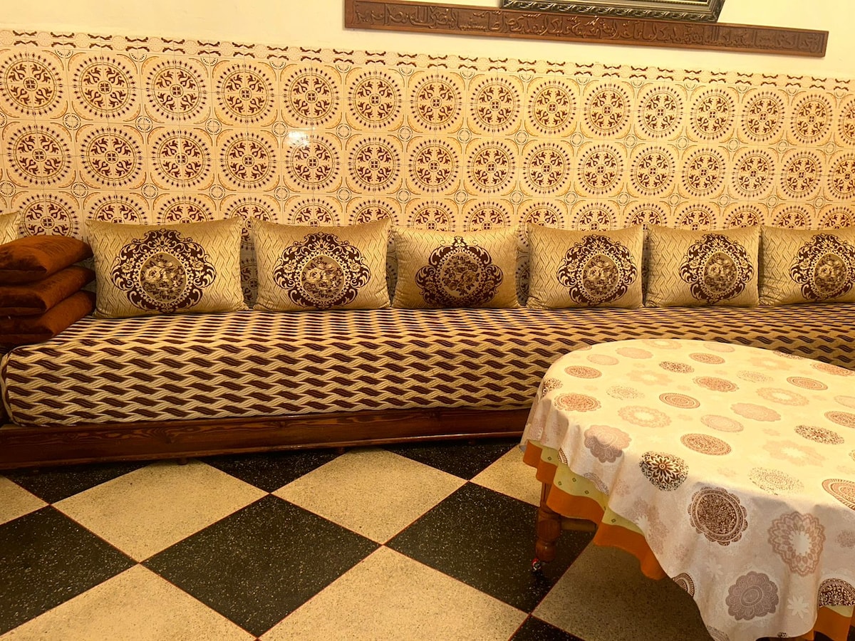 A traditional seating area features a patterned sofa with decorative cushions, backed by ornate tiles. The tiled floor displays a classic black and white checkerboard design, while a round table draped with a patterned tablecloth occupies the space, offering a blend of comfort and cultural style.