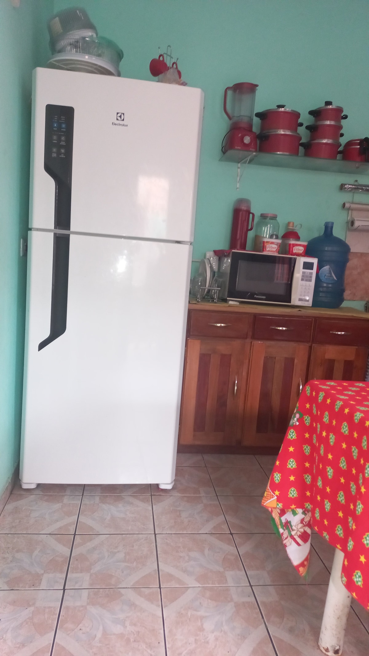 A compact kitchen area features a tall, white refrigerator next to a wooden cabinet with multiple drawers. On the counters, various kitchen appliances, including a microwave and a few decorative items, are visible. The walls are painted in a soft teal shade, complemented by tiled flooring.
