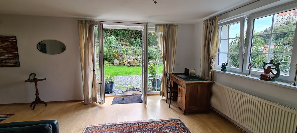 A bright and airy living space is depicted, featuring large glass doors that open to a lush garden. A wooden desk is placed against the wall, while a small side table is positioned nearby. Natural light floods the room, highlighting the wooden flooring.