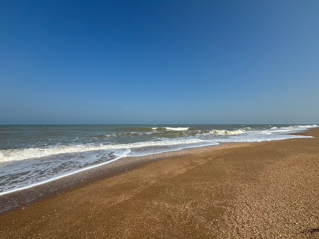 The image captures a wide stretch of sandy beach meeting the gentle waves of the sea under a clear blue sky. The shoreline is defined by a soft, undulating sand pattern, with foamy waves lapping at the edge.