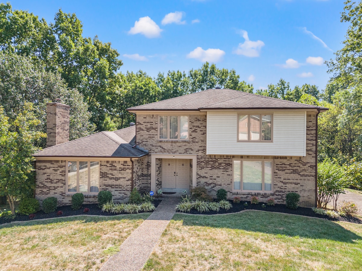The home's exterior is characterized by a two-story brick facade, surrounded by well-maintained landscaping. A paved pathway leads to the front entrance, framed by large windows that allow natural light to illuminate the interior. Lush greenery is visible in the background.