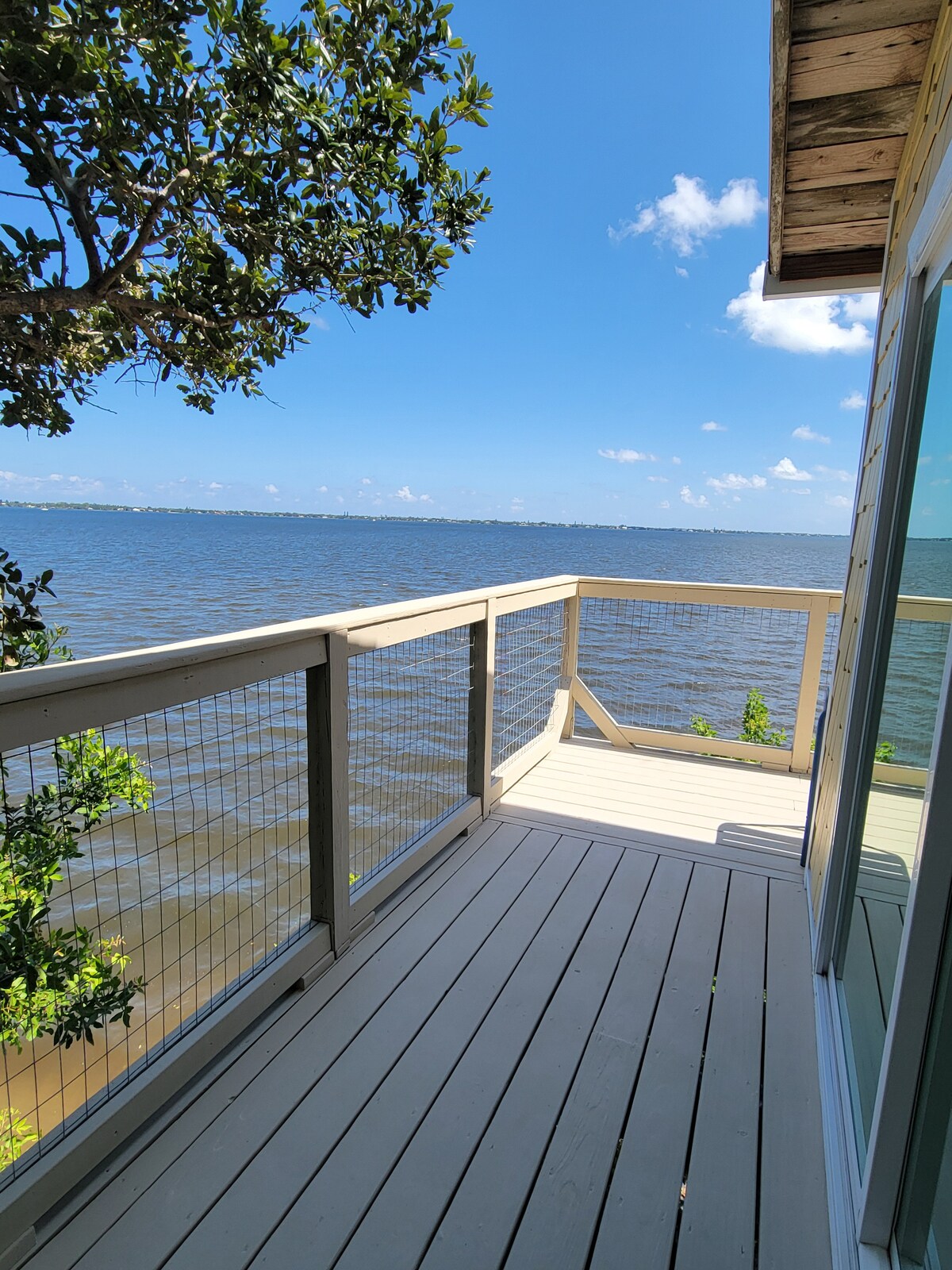 A wooden deck extends out over the water, offering views of the river. Bright blue skies and scattered clouds create a serene backdrop. Green foliage from trees frames the outdoor space, enhancing the connection to nature.