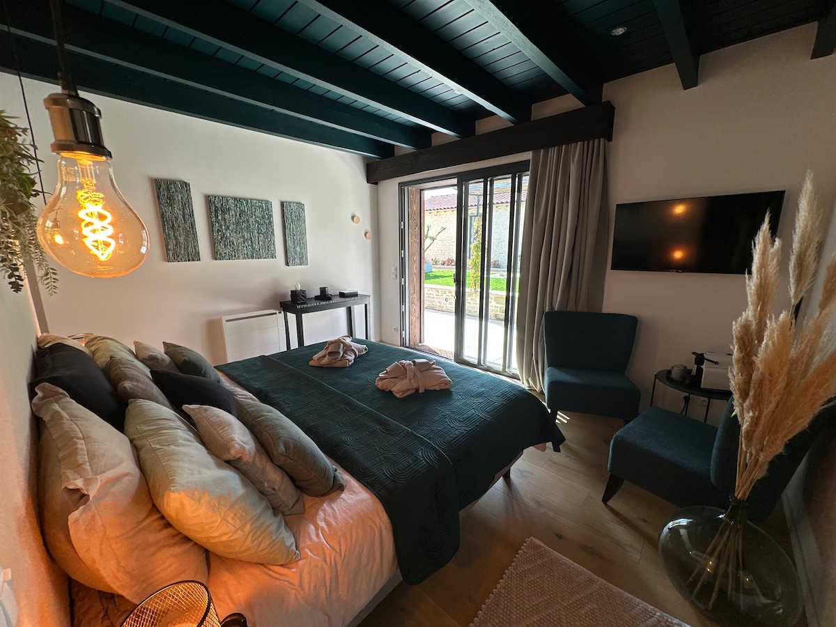 A well-appointed bedroom features a spacious bed with layered pillows and a dark comforter. A seating area is present, complemented by a decorative plant and a glass table. Large windows offer views of the outdoor garden, with natural light flowing into the space.