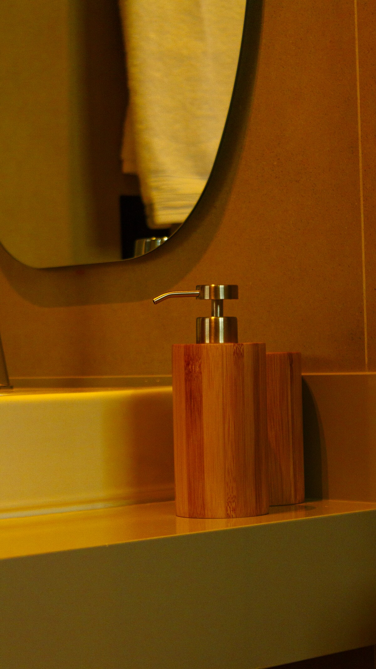 A modern bathroom countertop is shown, featuring a bamboo soap dispenser positioned beside a sink. The mirror above reflects soft light, enhancing the warm tones of the surrounding walls. Towels are neatly arranged in the background.