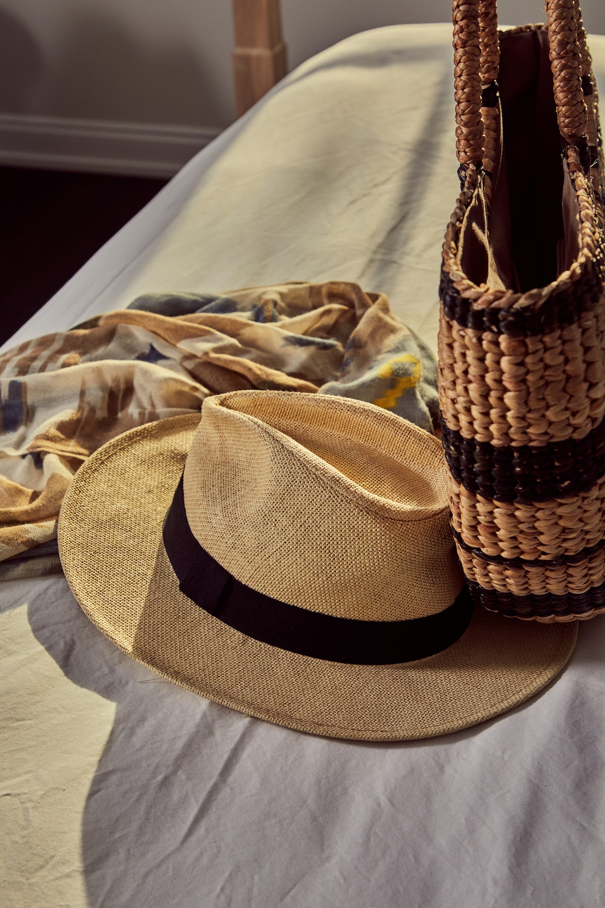 A stylish straw hat is placed on a light-colored bedspread alongside a woven bag and a lightweight scarf. The natural textures and warm tones create an inviting scene, suggesting comfort and relaxation.