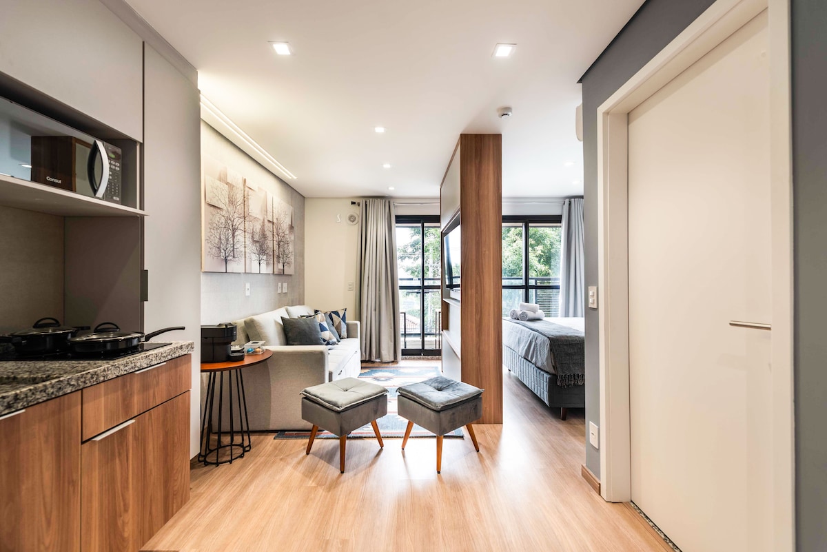 A modern apartment interior is showcased, featuring an open-plan layout. A kitchen area with a granite countertop is visible, accompanied by a small dining space. The living area contains a comfortable sofa and two poufs, with large windows providing natural light and views of greenery.