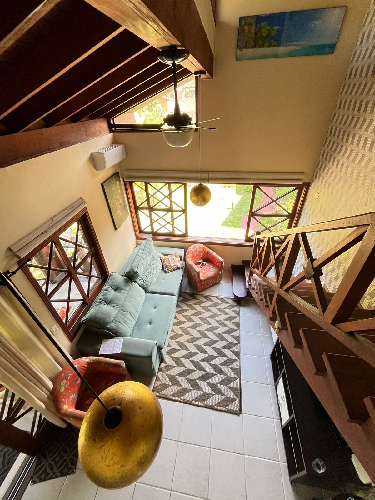 The image captures an upper view of a living area featuring a light blue couch and two vibrant chairs. A patterned rug covers the tiled floor, and large windows allow natural light to illuminate the space. Decorative items are present on the walls, enhancing the environment.