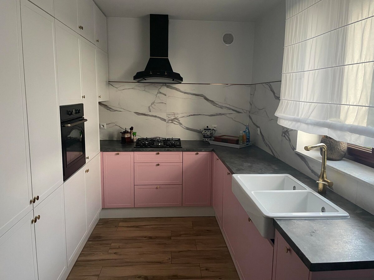 A modern kitchen features cabinetry with a combination of white and soft pink finishes. Marble-patterned backsplashes contribute to a sophisticated look. Equipped with a gas stove and an oven, the space is complemented by a large, farmhouse-style sink under a light-filtering window.