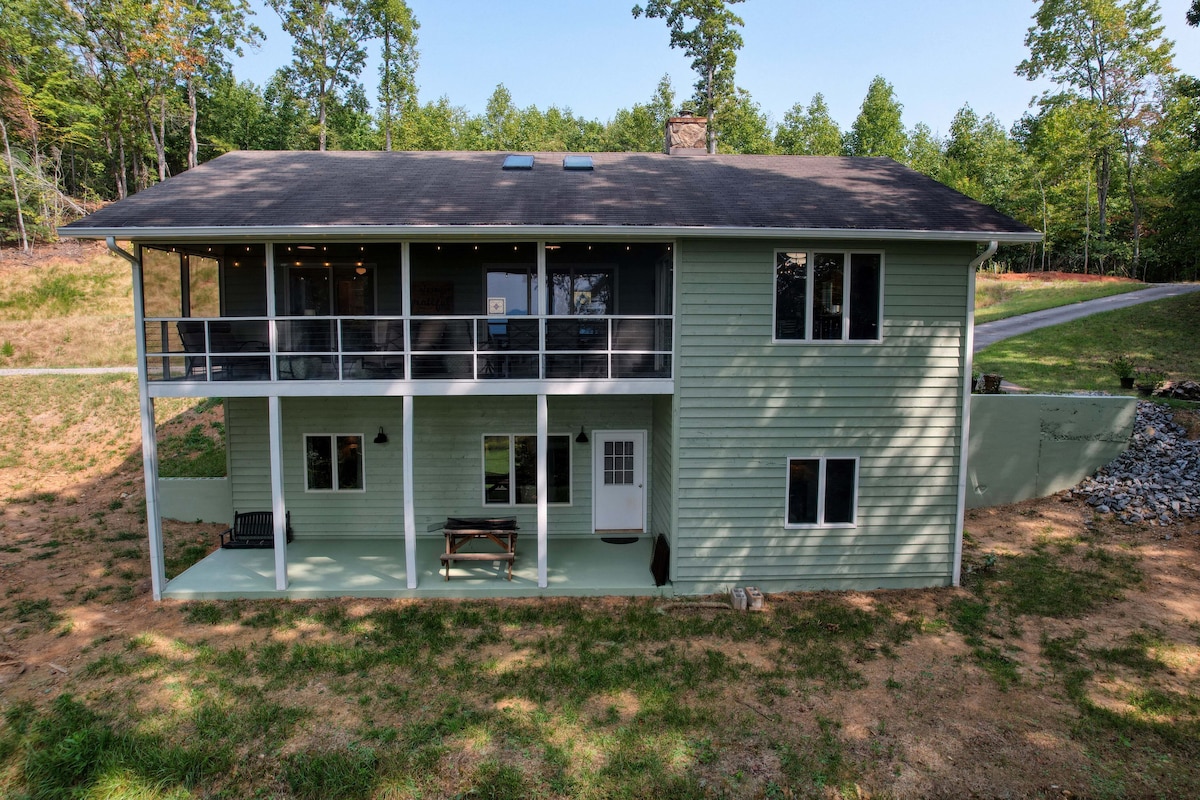 The exterior of a two-story light green house is captured amidst a lush forest setting. Large windows allow for ample natural light. A spacious porch features railings and is furnished with bench seating. A grassy area surrounds the home, offering a peaceful atmosphere.