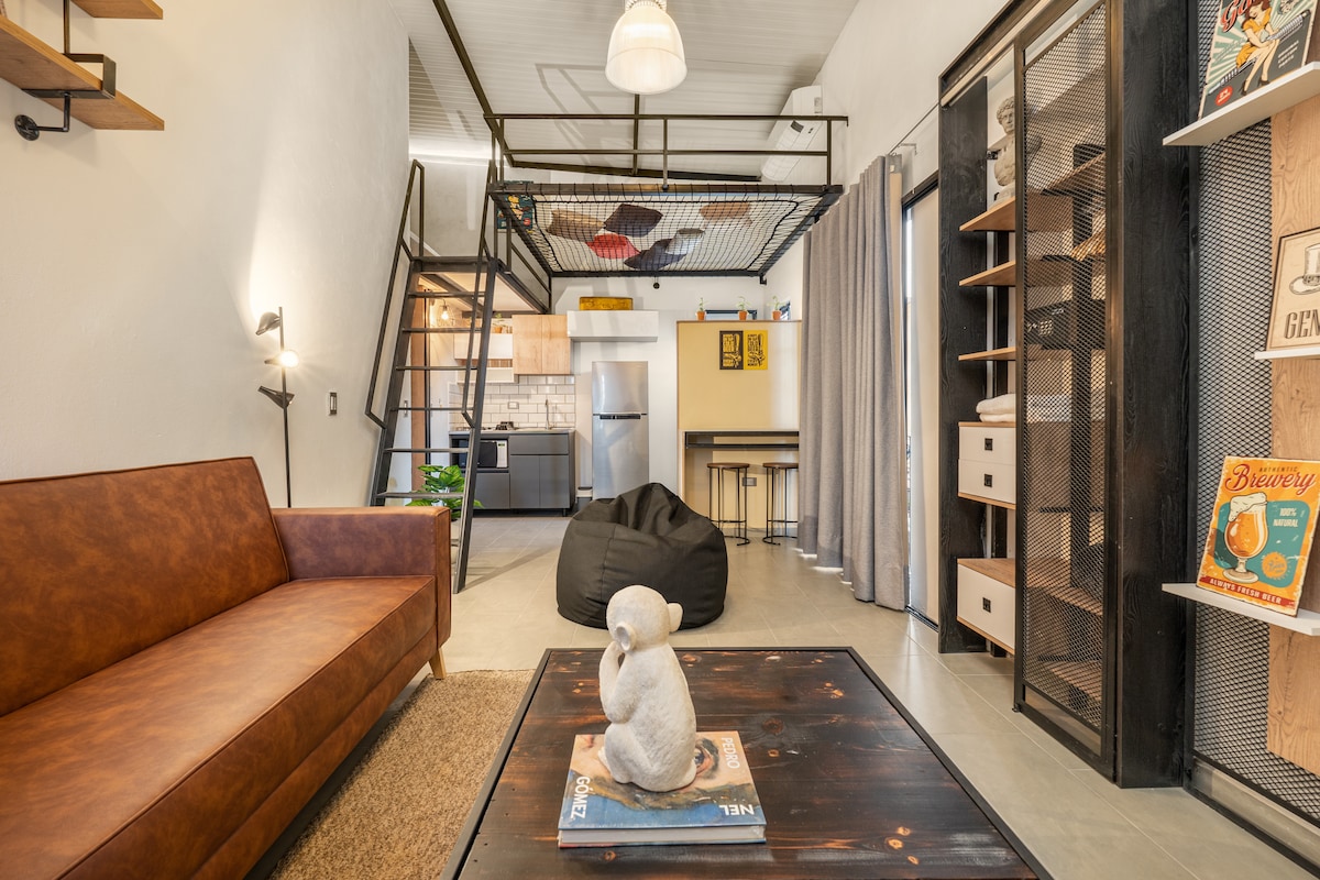 An open living area features a modern brown sofa on a woven rug, complemented by a decorative table with a small sculpture. A lofted sleeping area with metal railing and a fully equipped kitchen are visible in the background, along with shelves displaying colorful art and books.