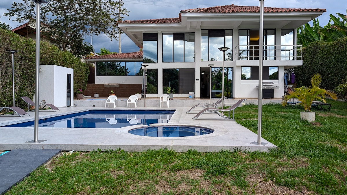 An inviting outdoor area features a sparkling pool accompanied by a jacuzzi. Lounge chairs are arranged around the water, with a landscaped garden and trees in the background. A modern house with large windows and a patio can be seen, creating a harmonious indoor-outdoor connection.