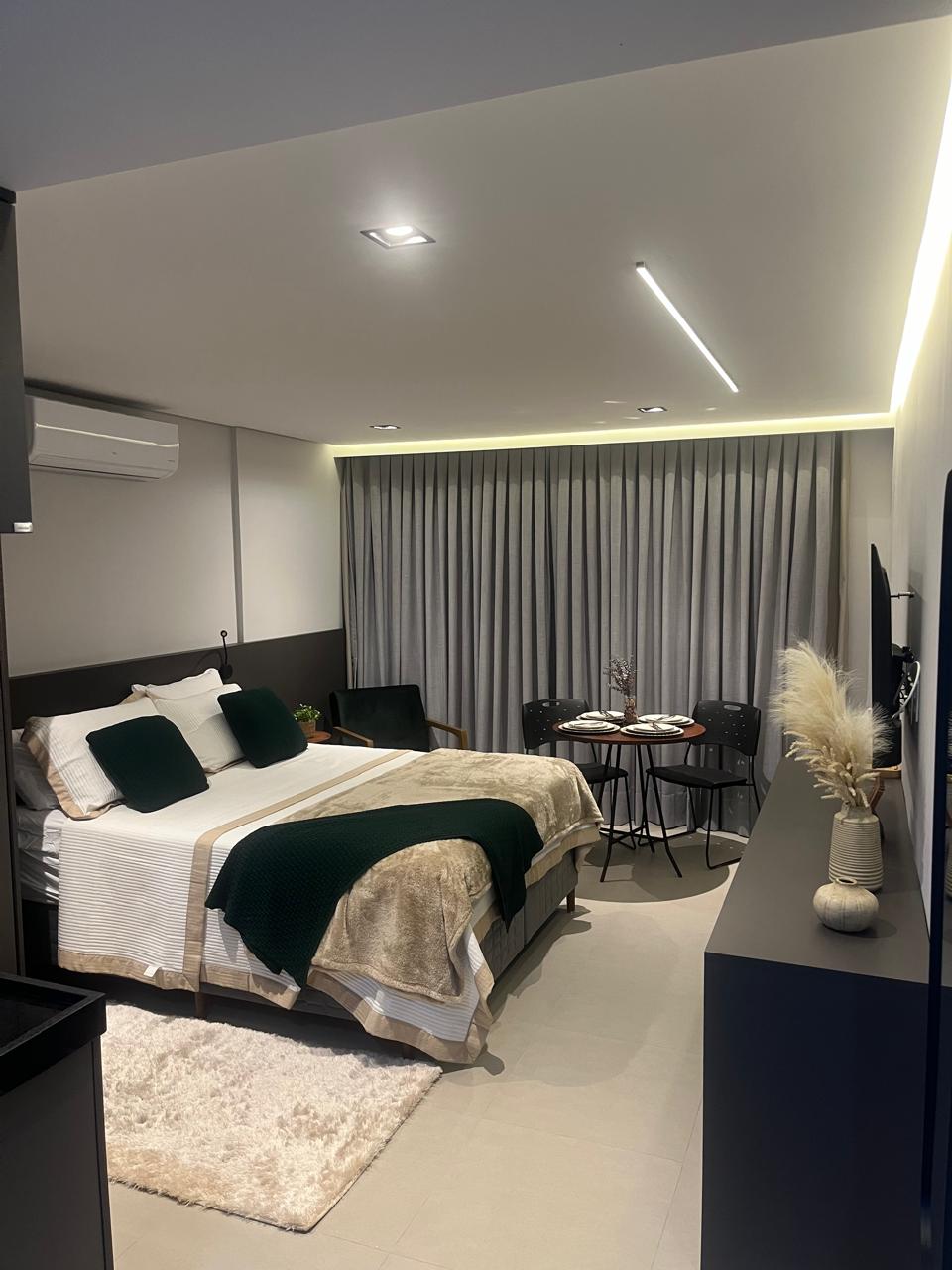 A well-appointed studio features a spacious bed adorned with dark green and neutral-toned linens. A small dining area with two chairs and a round table is positioned beside a large window, partially covered by light gray curtains. Soft lighting subtly highlights the modern decor.