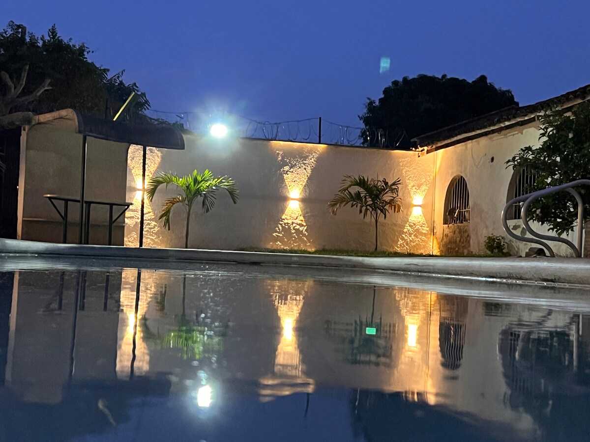 The tranquil pool area is illuminated by soft lights, creating reflections on the water's surface. A landscaped wall is adorned with greenery, while a shaded seating area is visible nearby. The serene ambiance suggests a peaceful evening atmosphere.