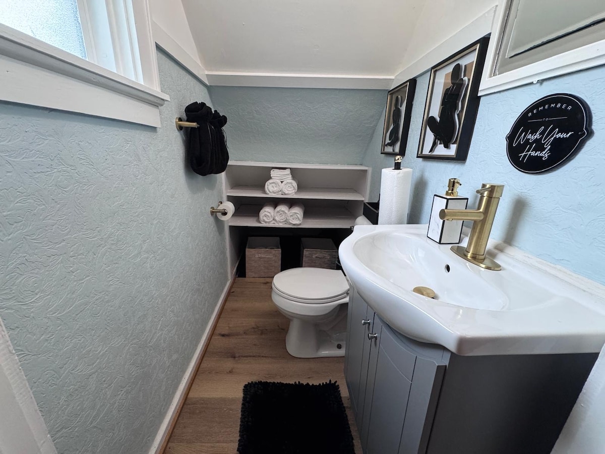 A well-appointed bathroom features a white vanity with a gold faucet beside a wall-mounted mirror. Neatly arranged towels are displayed on a shelf, while a black rug complements the space. Light blue walls provide a calming backdrop, enhancing the overall ambiance.