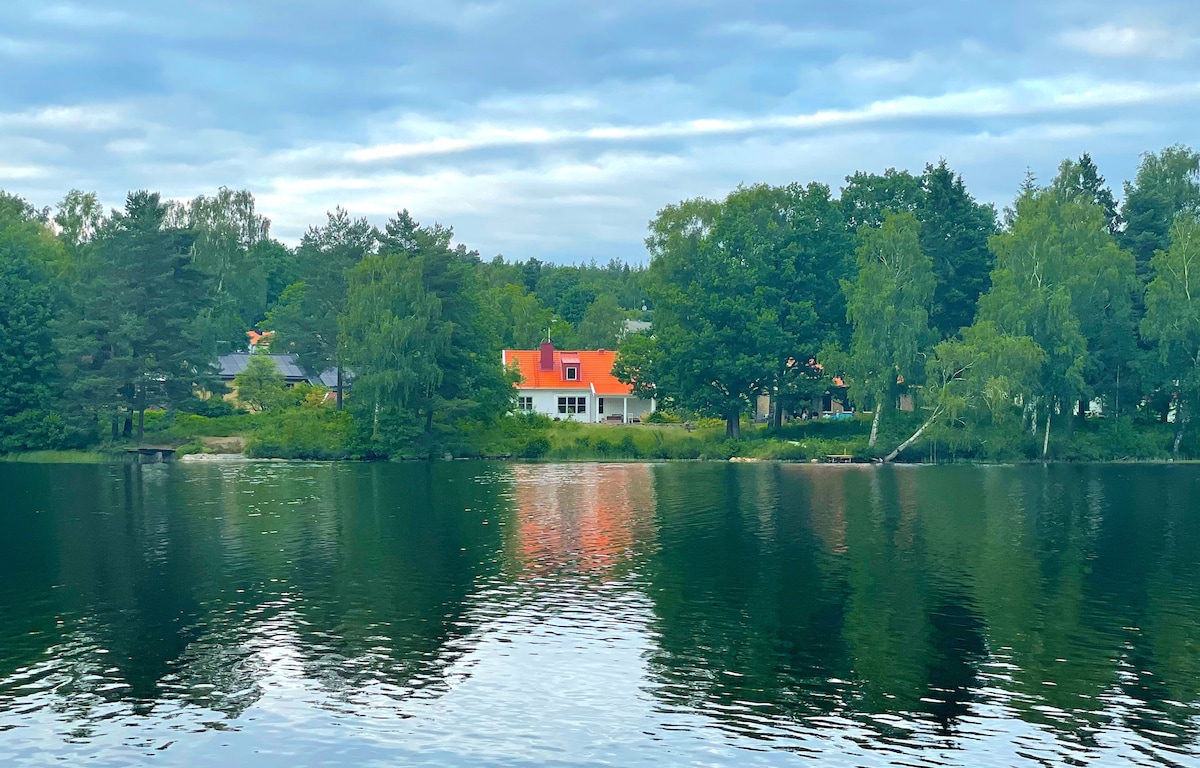 A lakeside view showcases a classic Swedish home with a vibrant orange roof. Surrounding trees reflect in the calm waters of the lake, creating a serene natural setting. The distant elements of the village can be seen peeking through the greenery.