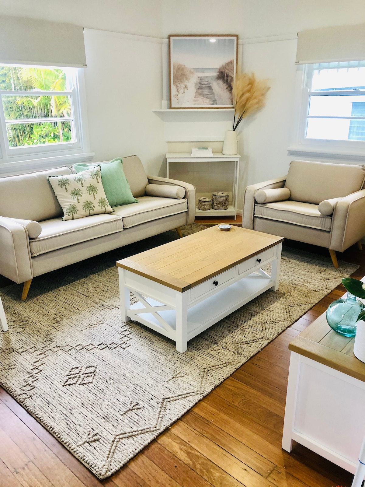 A cozy living area is furnished with light-colored, modern seating, featuring two armchairs and a comfortable sofa. A wooden coffee table sits at the center, complemented by a natural fiber rug. Natural light filters through the windows, enhancing the room's inviting ambiance.