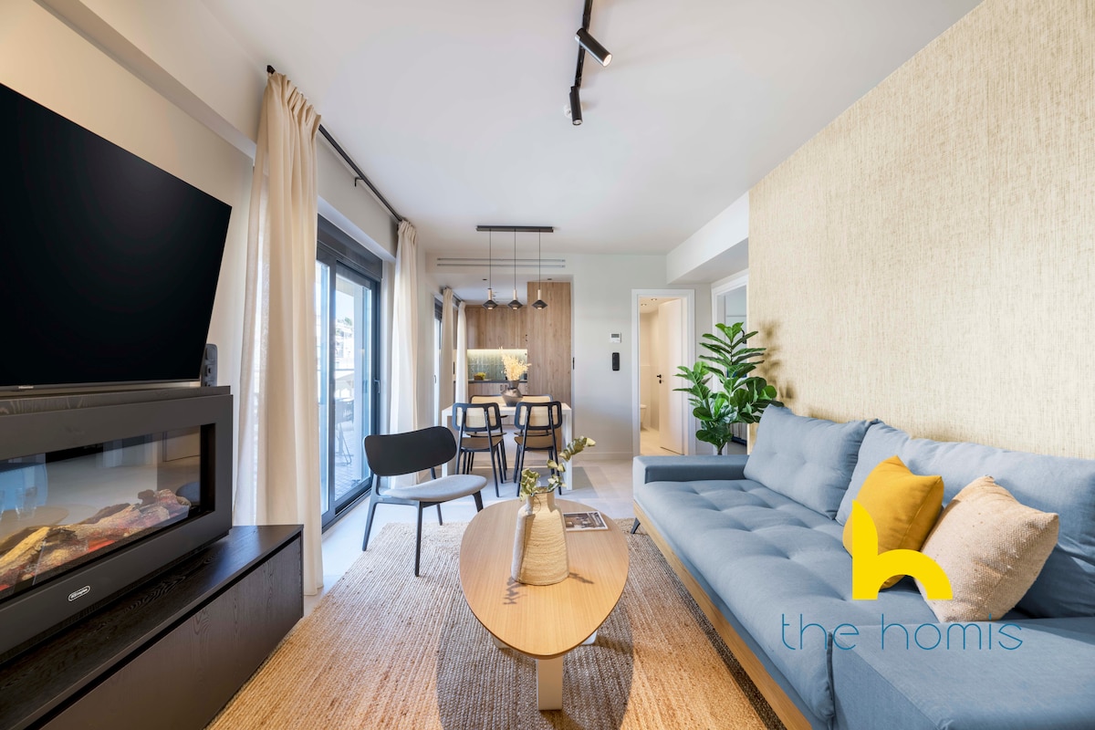 A cozy living room features a custom-made blue sofa positioned across from a flat-screen TV. Natural light streams through sliding glass doors that open to a serene patio. A round coffee table sits in front, complemented by a neutral-toned area rug and greenery for a calming touch.
