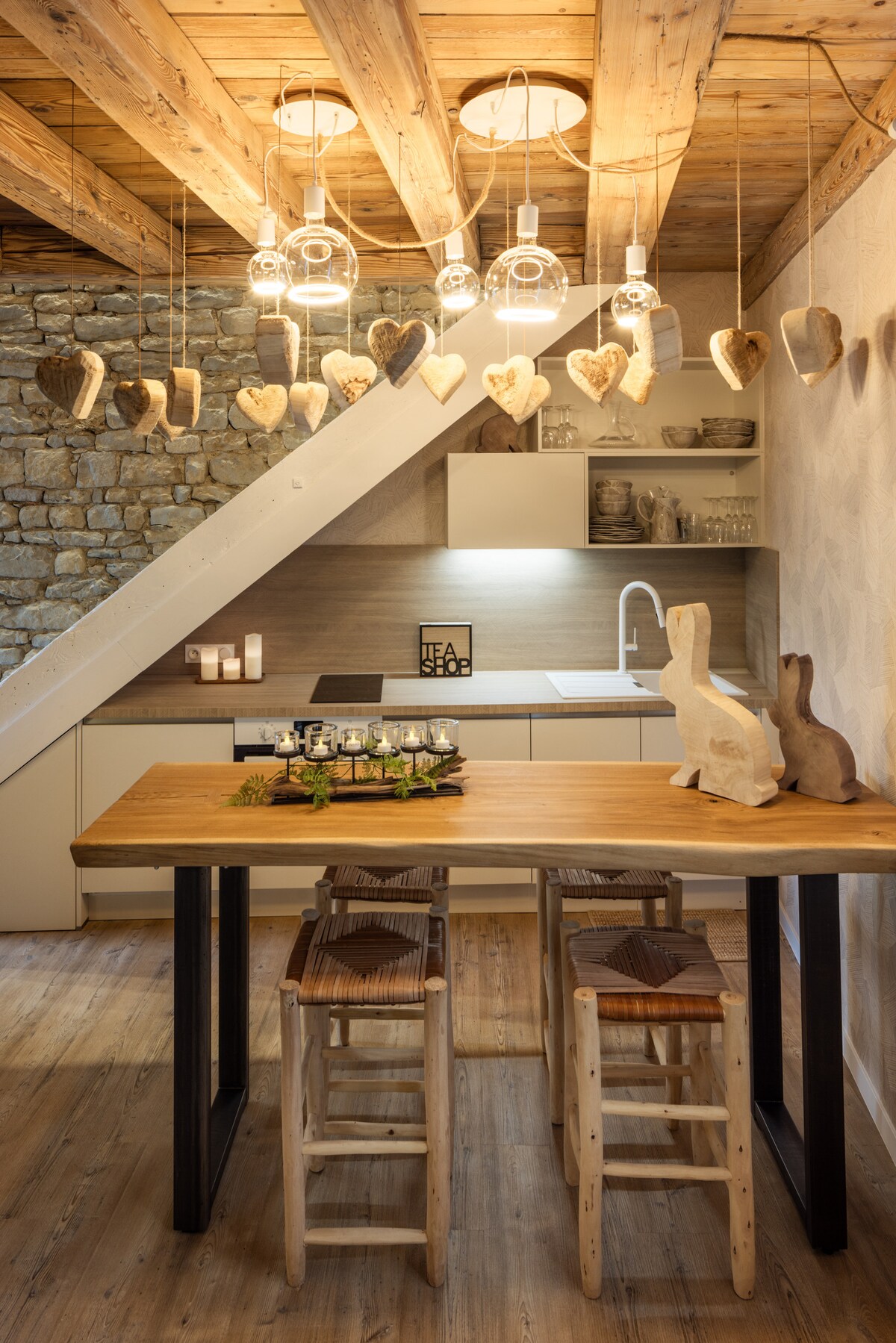 A well-appointed kitchen area features wooden beams and a rustic table with two stools. Heart-shaped decorations hang above, and natural materials are used throughout. Soft lighting enhances the inviting atmosphere, while a modern sink and cabinetry integrate contemporary design with traditional elements.