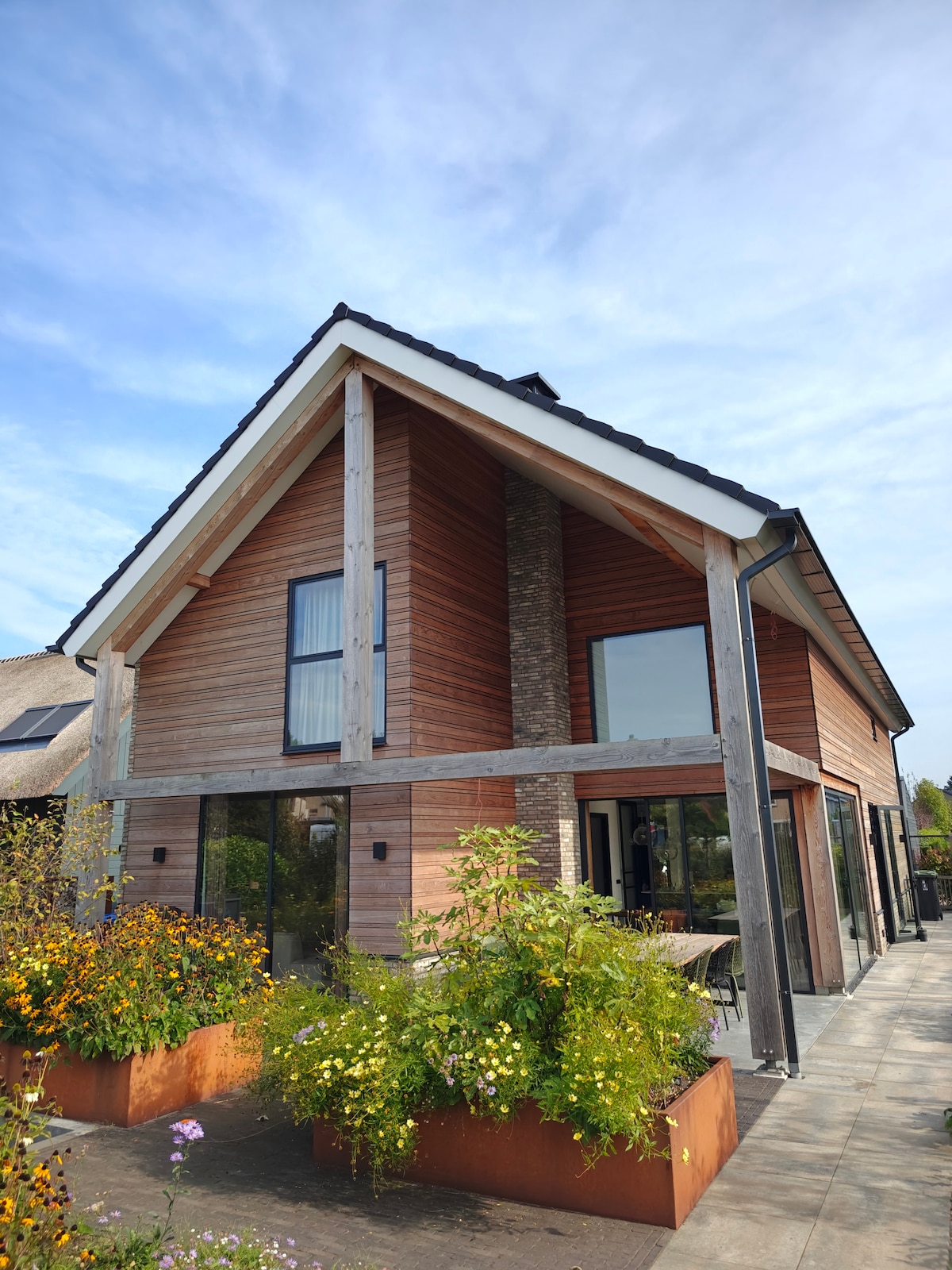 The exterior of a modern family home is highlighted, showcasing a blend of wood and stone materials. Large windows allow ample natural light, while vibrant flower boxes enhance the front. A spacious patio area is visible, providing an inviting outdoor space.