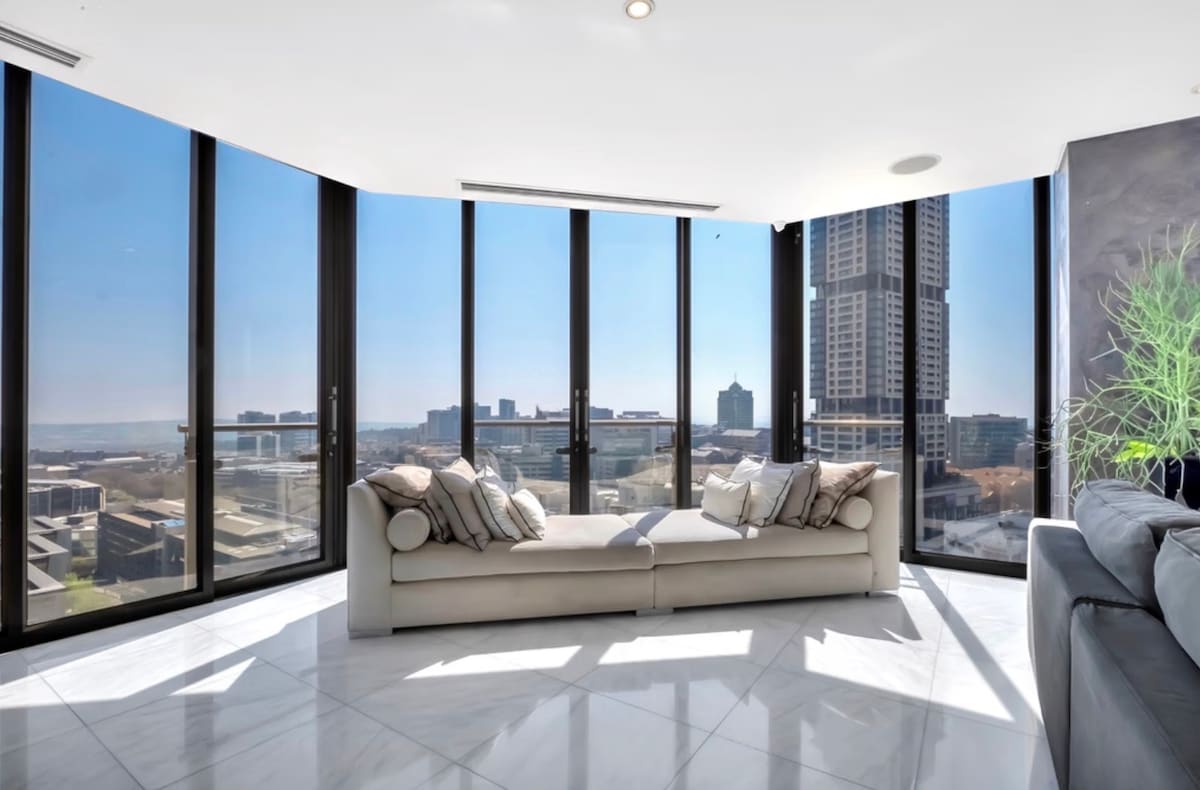 A bright and spacious living area features large floor-to-ceiling windows that offer panoramic views of the city skyline. The modern sofas, arranged for comfort, are accented with decorative cushions. Natural light fills the space, highlighting the sleek marble flooring.